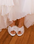 White sandals with large bows worn by a person in a white dress on a wooden floor.