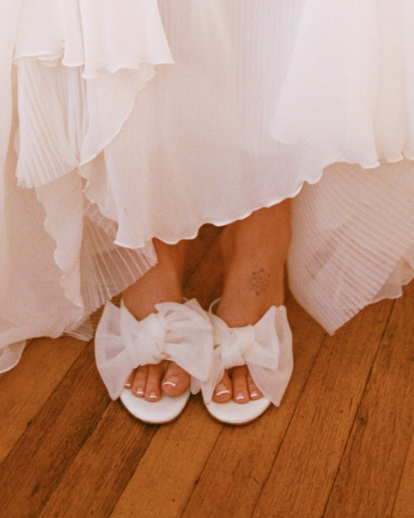 White sandals with large bows worn by a person in a white dress on a wooden floor.