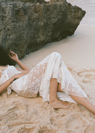 Woman in a white dress lying on a sandy beach with a large rock and ocean in the background