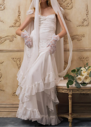 Woman in a white wedding dress with a veil and gloves, standing next to a table with flowers against an ornate wall.