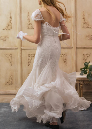 Woman in a white lace dress standing in a room with ornate wallpaper.