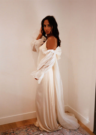 Woman in a white off-shoulder dress standing against a plain wall.