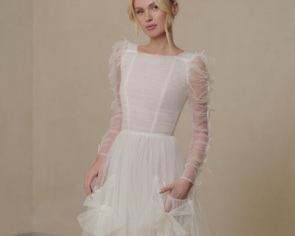 Woman wearing a white wedding dress with sheer sleeves in a room with beige walls and wooden floor.